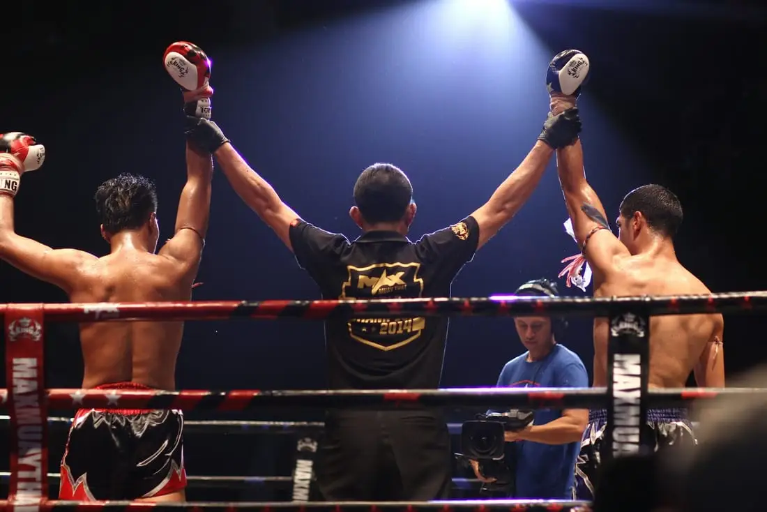 Sur un ring de muay thai éclairé, un arbitre lève les bras des deux combattants à l’issue du combat.