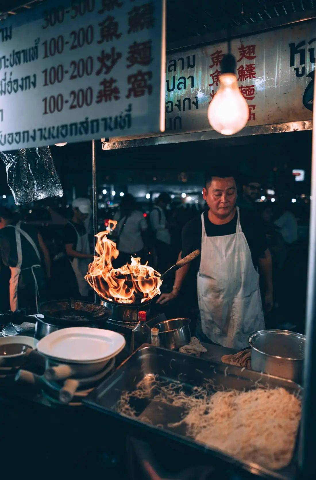 Un cuisinier fait flamber un wok sur un étal de rue éclairé à l’ampoule, de nuit.