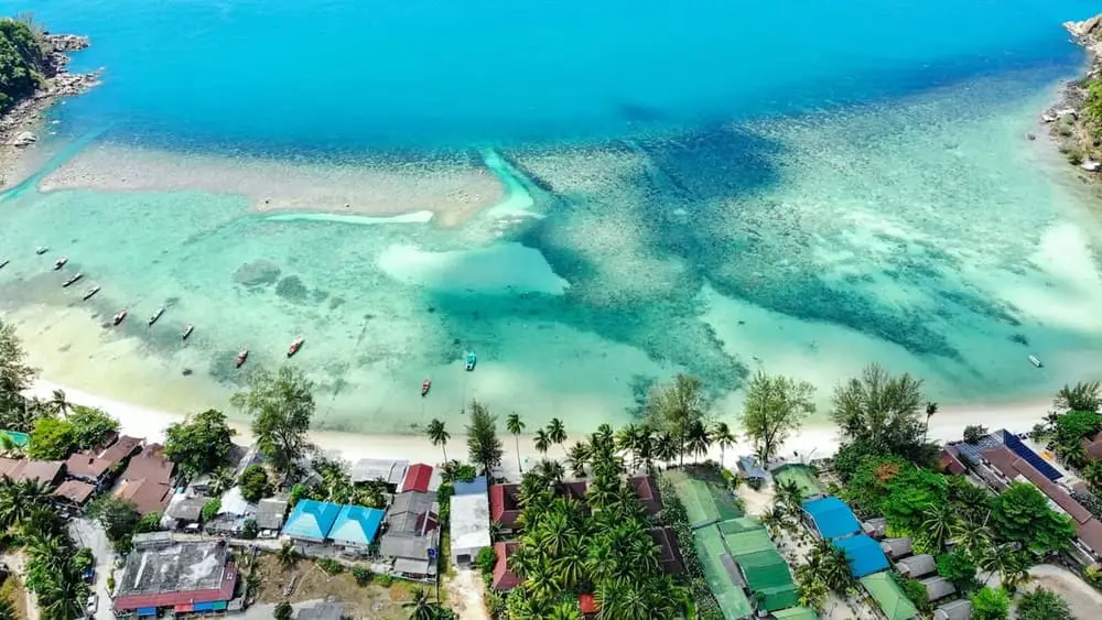Vue aérienne d’une baie de la mer d’Andaman : récif affleurant, eau turquoise et bateaux à longue queue mouillés le long de la plage.